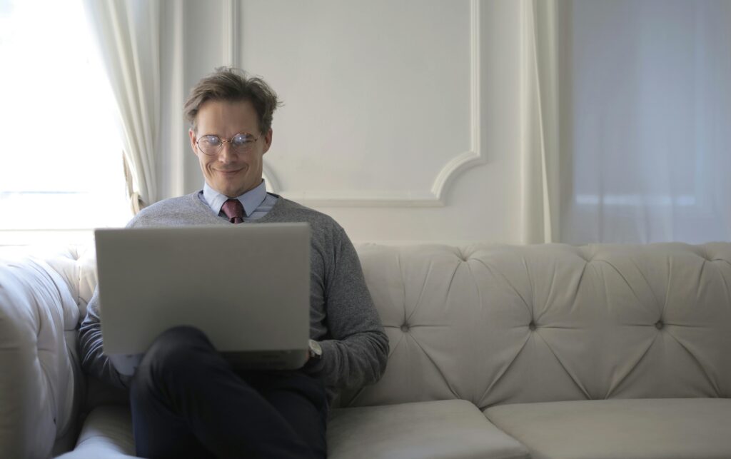 Man sitting on sofa using laptop, smiling and relaxed in a bright living room.