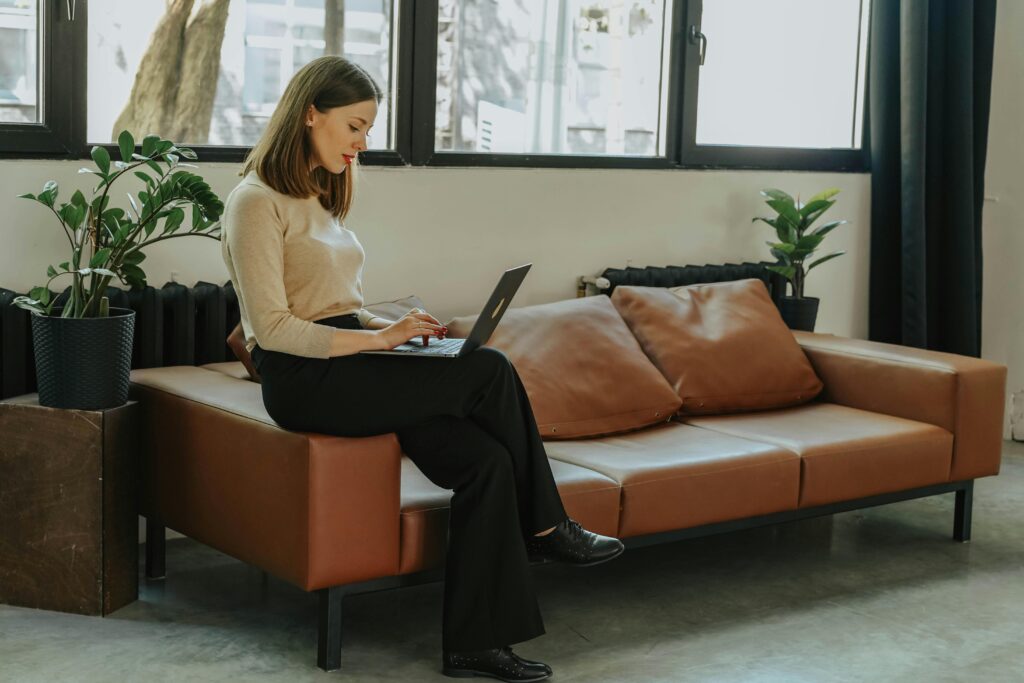 A professional woman working on a laptop, seated on a comfortable couch with indoor plants.